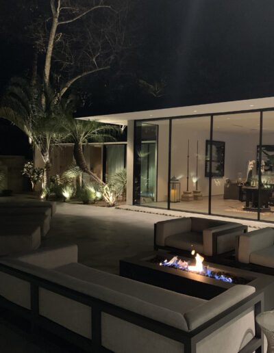 A modern outdoor patio with a fire pit, comfortable seating, and a view into a well-lit interior living space at night.